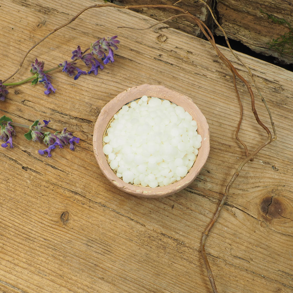 Weiße Lamecreme-Pellets in einer kleinen, rustikalen Schale, dekoriert mit lila Blüten und Zweigen, auf einem Holztisch.