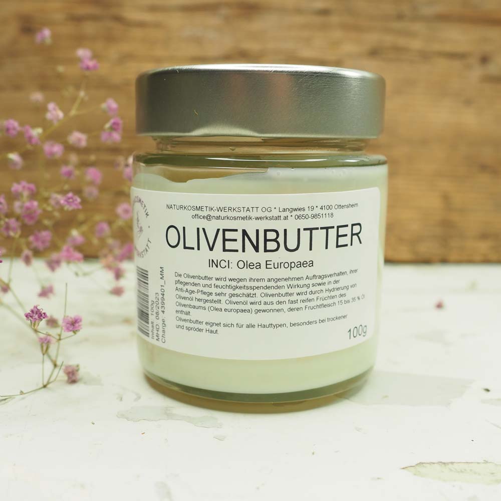 Glasbehälter mit Olivenbutter, Etikett sichtbar, dekoriert mit kleinen lila Blüten auf einem rustikalen Hintergrund